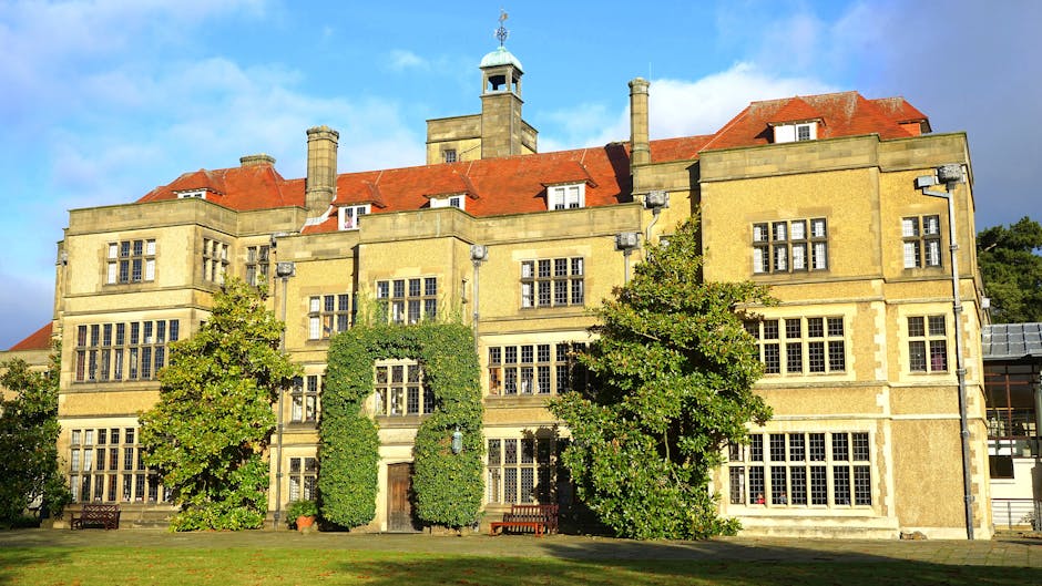 A large, historic-style house with a prominent tower and multiple chimneys, situated behind a neatly maintained garden with tall trees and shrubs. The property is partially obscured by a layer of fog